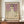 May include: Framed art print of Gustav Klimt's 1912 painting featuring a young woman in a white dress with floral embroidery against a purple floral background. The vintage-style poster has cream matting and dark wood frame, displayed on a wooden sideboard with a white ceramic vase and books in an elegant room with white paneled walls.