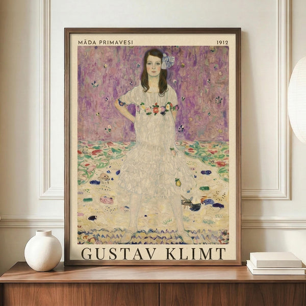 May include: Framed art print of Gustav Klimt's 1912 painting featuring a young woman in a white dress with floral embroidery against a purple floral background. The vintage-style poster has cream matting and dark wood frame, displayed on a wooden sideboard with a white ceramic vase and books in an elegant room with white paneled walls.