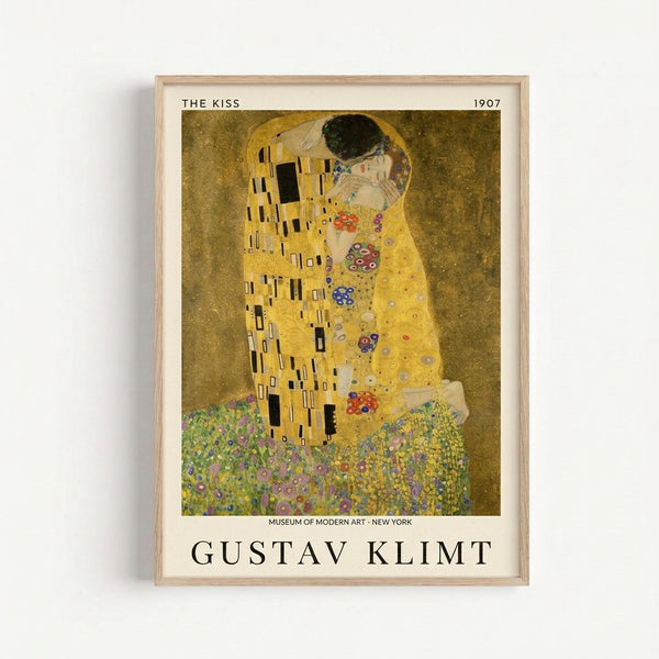 Framed art print of Gustav Klimt's famous painting "The Kiss" from 1907, showing two figures embracing in golden robes with geometric and floral patterns against a golden background with decorative wildflowers at the bottom. The poster includes museum attribution text and the artist's name in black lettering below the artwork. Light wood frame against white wall.