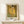 May include: Framed reproduction of Klimt's The Kiss artwork in golden wood frame, leaning against white wall on light wooden surface. Famous painting shows embracing couple in ornate gold and patterned robes against golden background with decorative florals below. Small wrapped white gift box with greenery sits in front of poster. Museum exhibition poster format with text at bottom.