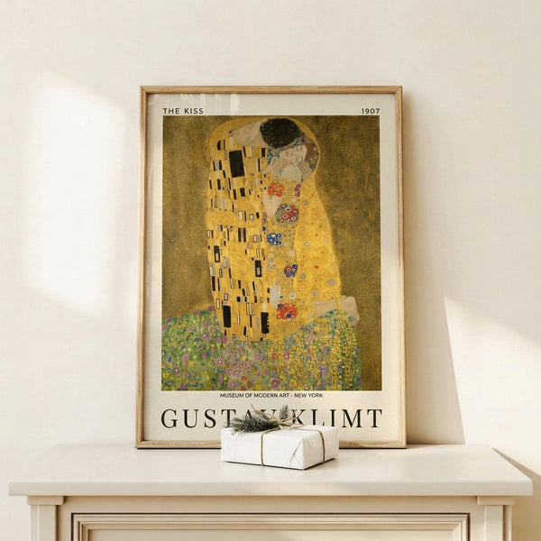 May include: Framed reproduction of Klimt's The Kiss artwork in golden wood frame, leaning against white wall on light wooden surface. Famous painting shows embracing couple in ornate gold and patterned robes against golden background with decorative florals below. Small wrapped white gift box with greenery sits in front of poster. Museum exhibition poster format with text at bottom.