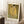 May include: Framed art print of Klimt's famous golden painting showing two embracing figures in ornate robes against a gold background with colorful floral patterns below. The beige-framed poster leans against white paneled walls on a white textured rug, with a coffee cup on a wooden block nearby and neutral fabric visible at the edge.