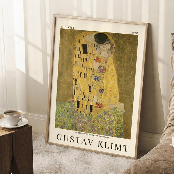 May include: Framed art print of Klimt's famous golden painting showing two embracing figures in ornate robes against a gold background with colorful floral patterns below. The beige-framed poster leans against white paneled walls on a white textured rug, with a coffee cup on a wooden block nearby and neutral fabric visible at the edge.