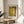 May include: A framed art print of Klimt's famous golden painting showing two embracing figures in ornate robes, displayed on a round wooden side table next to a beige upholstered chair. Soft natural light creates gentle shadows across the neutral-toned room setting with a plant visible in the corner.