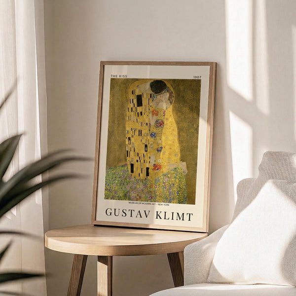 May include: A framed art print of Klimt's famous golden painting showing two embracing figures in ornate robes, displayed on a round wooden side table next to a beige upholstered chair. Soft natural light creates gentle shadows across the neutral-toned room setting with a plant visible in the corner.