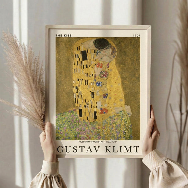 May include: Person in cream ruffled sleeves holding framed art print of Gustav Klimt's "The Kiss" from 1907. Golden artwork shows embracing couple in ornate robes against decorative background. Light wood frame with museum text. Dried pampas grass held in other hand. Neutral curtained background with soft natural lighting.