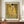 May include: Framed reproduction of Klimt's famous painting showing two embracing figures in golden robes with geometric and floral patterns against a shimmering gold background. The museum-style poster sits on a wooden credenza beside a white ceramic vase and books in a neutral-toned room with white walls and molding.