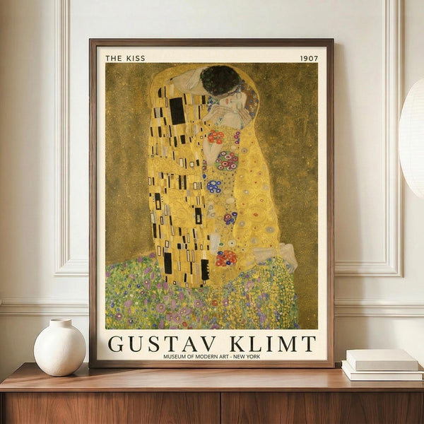 May include: Framed reproduction of Klimt's famous painting showing two embracing figures in golden robes with geometric and floral patterns against a shimmering gold background. The museum-style poster sits on a wooden credenza beside a white ceramic vase and books in a neutral-toned room with white walls and molding.