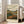 May include: Framed vintage-style landscape poster leaning against beige couch in bright living room with white walls and large windows. Print features rolling hills, winding river, mountains, and industrial buildings in earthy greens, oranges, and browns with cloudy sky. Text reads "HAND FLORA OFFSET" at bottom. Light wood frame on hardwood floors.