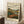 May include: Framed vintage-style landscape poster leaning against white wall in cozy room setting. Artwork shows rolling hills in earth tones with winding river, distant industrial buildings, and cloudy sky in lithograph print style. Light wood frame, displayed on white textured rug beside wooden side table with coffee cup and blanket corner visible.