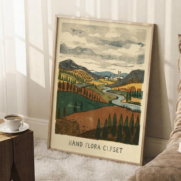 May include: Framed vintage-style landscape poster leaning against white wall in cozy room setting. Artwork shows rolling hills in earth tones with winding river, distant industrial buildings, and cloudy sky in lithograph print style. Light wood frame, displayed on white textured rug beside wooden side table with coffee cup and blanket corner visible.