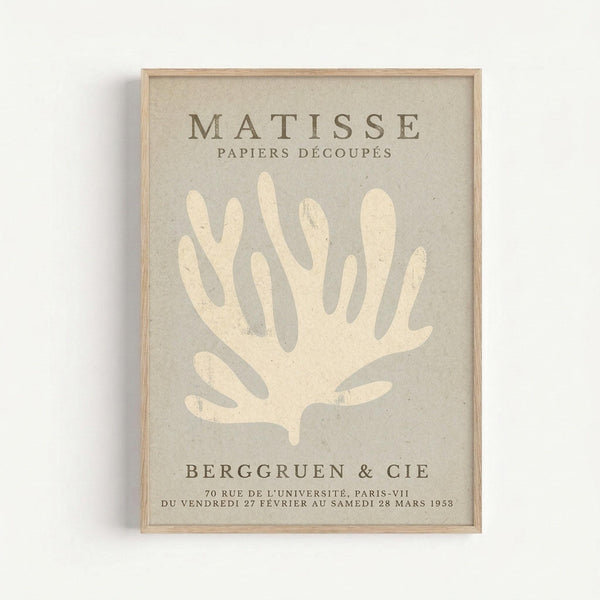 Framed poster featuring a cream-colored coral or seaweed silhouette on beige textured background. Text reads "MATISSE PAPIERS DÉCOUPÉS" at top and "BERGGRUEN & CIE" with French gallery details at bottom. Natural wood frame against white wall creates minimal shadow.
