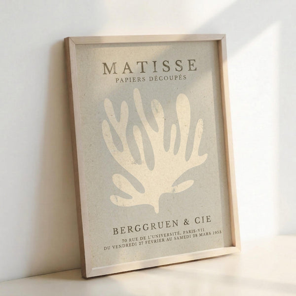 A framed exhibition poster featuring a stylized white coral or seaweed form against a soft gray-green background. The minimalist artwork shows "MATISSE" and "PAPIERS DÉCOUPÉS" at the top, with "BERGGRUEN & CIE" and French gallery text at the bottom. The poster is displayed in a light wooden frame, leaning against a white wall with natural lighting creating subtle shadows.
