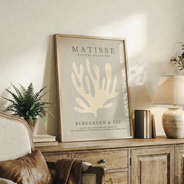 May include: Framed beige and cream botanical poster featuring white coral-like plant form, displayed on rustic wood sideboard in neutral living room with fern plant, ceramic vase, books, and beige upholstered chair with brown leather throw pillow