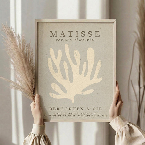 May include: Person in cream sweater holding framed vintage-style art poster featuring white coral or seaweed design on beige background, with dried pampas grass stems in other hand, neutral curtained window backdrop