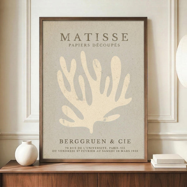 May include: Framed Matisse exhibition poster with cream coral silhouette on sage green background, displayed on wooden credenza beside white ceramic vase and stacked books in elegant room with beige walls and white molding
