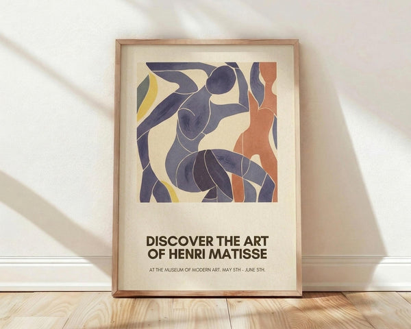 May include: Framed poster with abstract figures in blue, orange and yellow cut-out style artwork displayed against white textured wall on light wood floor. Features dancing forms in Matisse's signature paper collage aesthetic with exhibition text below reading "Discover the Art of Henri Matisse" and museum details. Natural window light creates soft shadows across the minimalist interior setting.