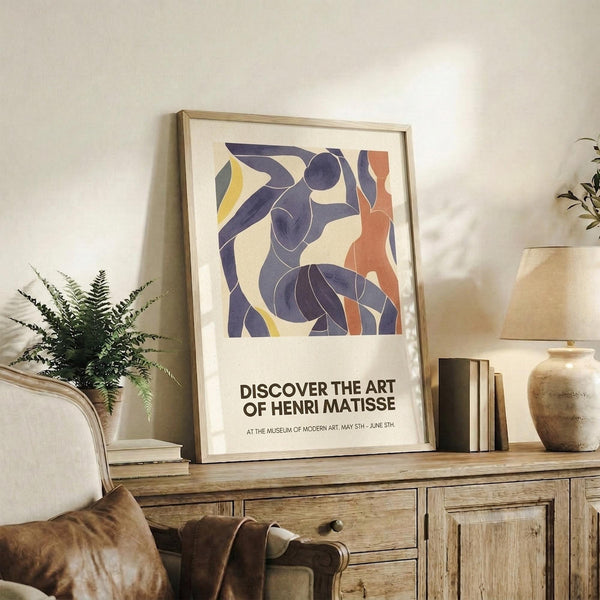 May include: Framed art print featuring abstract blue figures in Matisse's cut-out style with orange and yellow accents, displayed on rustic wood credenza alongside fern plant, ceramic vase, and books in warm-toned living room setting with cream upholstered chair