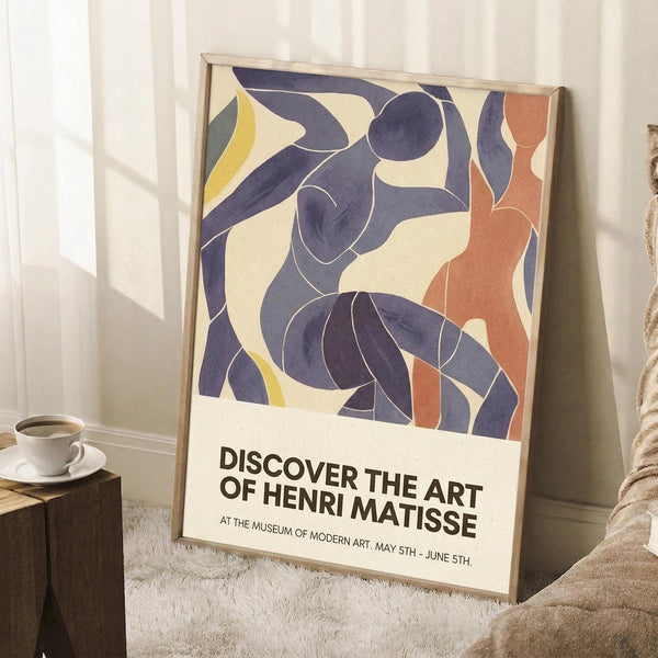 May include: Framed poster with abstract blue cut-out style figures dancing against cream background, with orange and yellow accents. Exhibition text reads "Discover the Art of Henri Matisse" at bottom. Light wood frame leaning against white wall corner next to wooden side table with white coffee cup and saucer, beige textured rug, and cream throw blanket visible in cozy interior setting with natural window light.