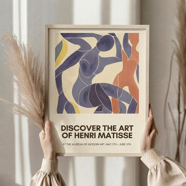 May include: Person holding framed poster featuring abstract cut-out style figures in deep blue, coral orange, and yellow against cream background. Modern minimalist artwork with flowing organic shapes reminiscent of dancers. Light wood frame, neutral beige clothing, dried pampas grass visible to left, soft natural lighting against white curtained window.
