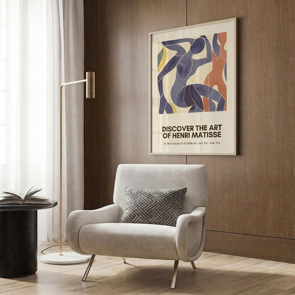 May include: Framed Henri Matisse exhibition poster featuring abstract blue figures in cut-out style with orange and yellow accents, displayed above a beige upholstered armchair with geometric pillow in modern living room with wood paneling, brass floor lamp, and side table with open book.