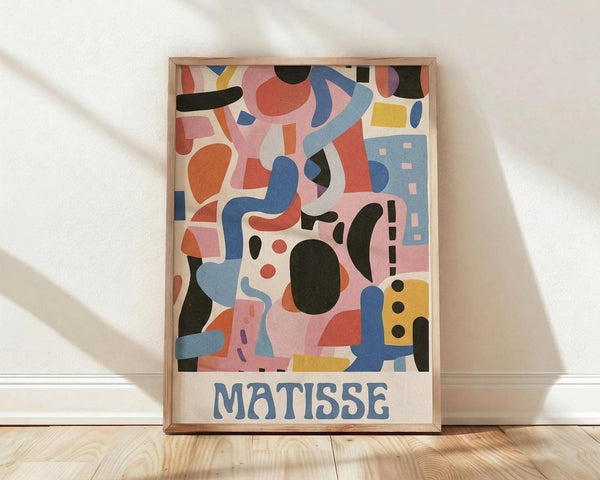 May include: Framed art print leaning against white wall on light wood floor with natural shadows. Abstract colorful composition featuring organic shapes in coral pink, blue, yellow, black, and orange. Modern minimalist interior setting with white baseboard trim. Natural wood frame surrounds poster with "MATISSE" text at bottom in blue letters.