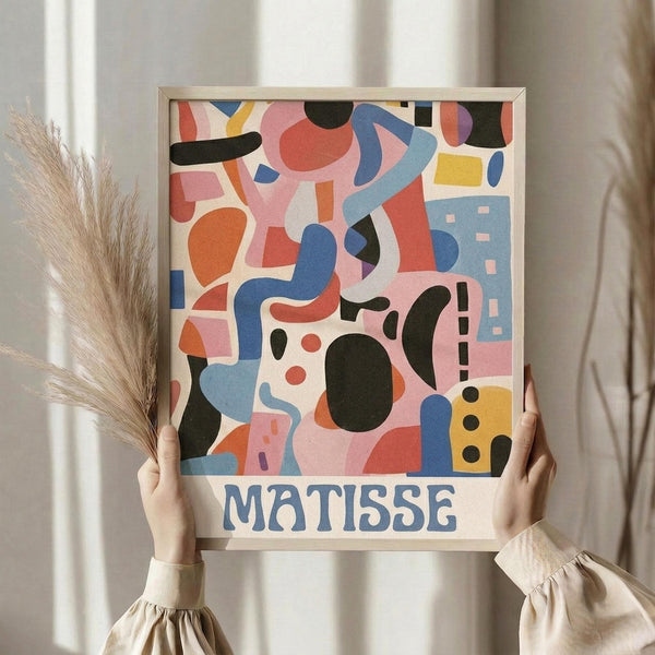 May include: Person holding framed abstract art poster with colorful geometric shapes in pink, blue, orange, yellow, and black cut-out style, with "MATISSE" text below artwork. Light wood frame, neutral beige clothing, dried pampas grass visible, soft natural lighting against cream curtained background.