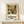May include: Framed botanical art print displayed on light wood console table against cream wall. Artwork features stylized flowers and leaves in warm earth tones - golden yellow, sage green, brown, and cream. Print shows "DISCOVER THE ART OF HENRI MATISSE" text below collage-style floral design. Natural wood frame with cream matting. Small wrapped gift box with twine sits nearby on table surface.
