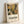 May include: A framed art print leaning against a white wall, featuring an abstract floral collage design with layered botanical elements in warm earth tones - golden yellow, sage green, brown, and cream. The artwork shows stylized flowers, leaves, and plant motifs in a cut-paper aesthetic. Below the colorful botanical illustration, black text reads "DISCOVER THE ART OF HENRI MATISSE" with smaller text indicating a museum exhibition. The print is displayed in a light wooden frame.