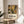 May include: Matisse-inspired botanical poster with stylized flowers and leaves in warm yellows, greens, and browns, displayed in light wood frame on round wooden side table next to beige upholstered chair. Soft natural lighting creates gentle shadows on neutral wall background with plant leaves visible in corner.