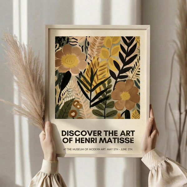 May include: Hands holding a framed botanical art print featuring abstract flowers and leaves in warm earth tones of brown, yellow, and green. The artwork shows layered floral collage elements in a modern style. Light wood frame against neutral curtained background with dried pampas grass visible to the left. Exhibition-style poster format with text below the botanical illustration.