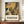 May include: Framed poster with botanical illustration in Matisse-inspired style featuring overlapping flowers and leaves in warm tones of yellow, brown, and green against cream background. Bold text below reads "DISCOVER THE ART OF HENRI MATISSE" with museum exhibition details. Display shows poster leaning on wooden sideboard next to white ceramic vase and stacked books in bright, modern interior with white walls and crown molding.