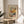 May include: Henri Matisse exhibition poster print in thin gold frame displayed on round wooden side table. Abstract floral artwork features vibrant yellow, orange, blue, green and white brushstrokes with "creativity takes courage" quote below. Soft natural lighting creates shadows across beige wall, with plant leaves and neutral upholstered chair visible in cozy interior setting.