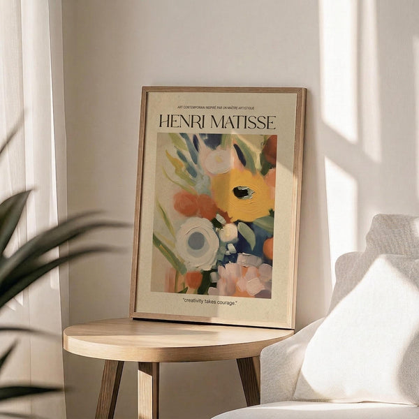 May include: Henri Matisse exhibition poster print in thin gold frame displayed on round wooden side table. Abstract floral artwork features vibrant yellow, orange, blue, green and white brushstrokes with "creativity takes courage" quote below. Soft natural lighting creates shadows across beige wall, with plant leaves and neutral upholstered chair visible in cozy interior setting.