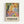 Colorful abstract art poster in light wood frame showing stylized fruits, flowers, and foliage in vibrant yellows, greens, oranges, and purples. Bold black tree branches frame organic shapes and botanical elements painted in loose, expressive brushstrokes. Text at bottom reads "DISCOVER THE ART OF HENRI MATISSE" with museum exhibition details. Poster mounted on white wall with soft shadow.