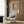 May include: Colorful Henri Matisse exhibition poster featuring vibrant fruits and flowers in yellow, orange, green, and blue tones, displayed in light wood frame on textured brown wall panel above beige upholstered armchair with geometric patterned pillow. Modern brass floor lamp and dark side table with open book complete the cozy reading nook scene with natural lighting from nearby window.