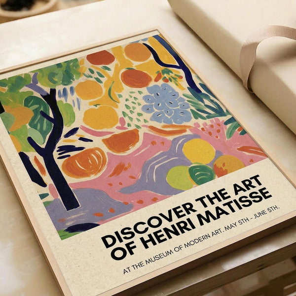 May include: Colorful Matisse-style exhibition poster featuring abstract fruit and flowers in vibrant yellows, oranges, blues, and greens with text "Discover the Art of Henri Matisse" displayed on white surface next to cream-colored lamp and neutral home decor