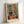 May include: Framed art print leaning against white wall showing colorful interior scene with black and white cat on wooden floor, open French doors, chair with blue cushion, potted plants, and vibrant orange, green, and yellow color palette in Matisse-inspired style. Light wood frame with white matting.