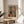 May include: Framed Henri Matisse art print displayed on round wooden side table next to beige upholstered chair. The colorful interior scene shows a room with orange walls, wooden floors, black and white cat, green chair, and windows. Soft natural lighting creates shadows across neutral walls. Plant leaves visible in corner of minimalist living space setting.