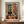 May include: Framed Henri Matisse-style interior painting showing colorful room with black and white cat sitting on orange wooden floor, green chair with pillow, dark French doors opening to garden view, vibrant orange and blue walls with botanical artwork. Print displayed on wooden credenza beside white ceramic vase and stacked books in neutral modern room with white walls and crown molding.