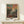 May include: Framed art print featuring an impressionistic landscape with vibrant orange and pink sky, lush green foliage with small orange fruits, and silhouetted figures beneath trees. The artwork displays thick, expressive brushstrokes in a post-impressionist style. Set in natural wood frame on light wooden surface with small white gift box and pine sprig against cream wall.