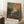 May include: Framed art print leaning against white wall corner showing colorful landscape painting with vibrant orange and green foliage, figures beneath fruit trees, and dramatic sky in blues, pinks and yellows. Natural wood frame, positioned on white textured rug next to wooden side table with coffee cup and blanket.