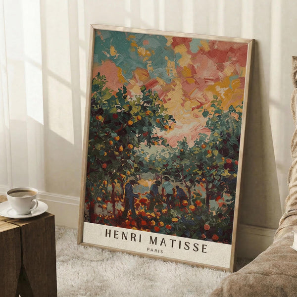 May include: Framed art print leaning against white wall corner showing colorful landscape painting with vibrant orange and green foliage, figures beneath fruit trees, and dramatic sky in blues, pinks and yellows. Natural wood frame, positioned on white textured rug next to wooden side table with coffee cup and blanket.