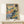 May include: Framed Matisse-style landscape poster with colorful abstract brushstrokes in blues, greens, oranges and yellows, showing trees, water and buildings. Natural wood frame sits on light wooden surface next to small wrapped gift box against cream wall. Vibrant Fauvism art style with loose, expressive painting technique.