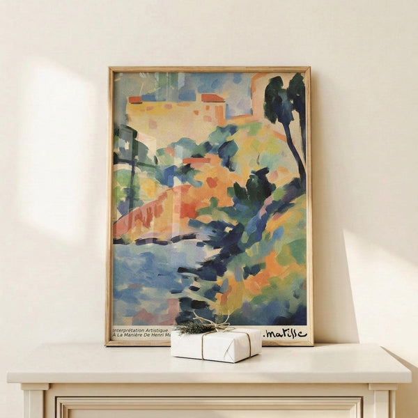 May include: Framed Matisse-style landscape poster with colorful abstract brushstrokes in blues, greens, oranges and yellows, showing trees, water and buildings. Natural wood frame sits on light wooden surface next to small wrapped gift box against cream wall. Vibrant Fauvism art style with loose, expressive painting technique.