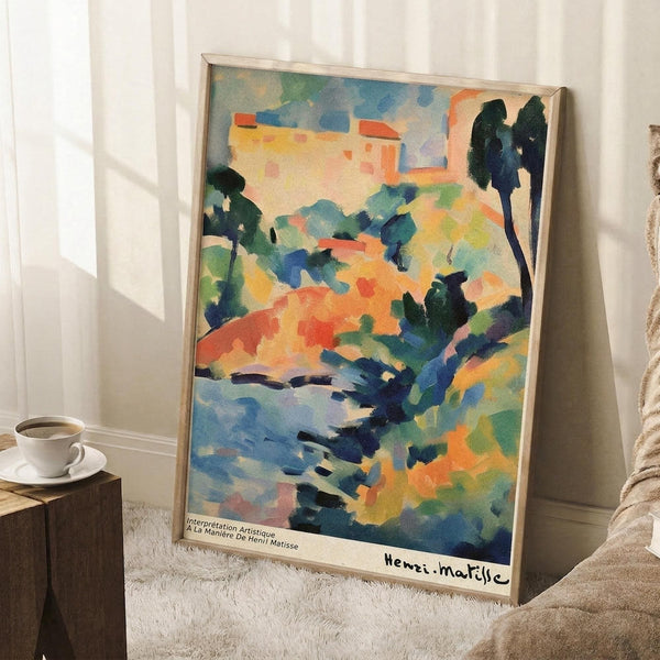 May include: Framed poster print featuring colorful abstract landscape with loose brushstrokes in blues, greens, oranges, and yellows depicting trees, buildings, and water. Light wood frame leaning against white wall on textured rug, with coffee cup on wooden stool nearby and cozy blanket visible.