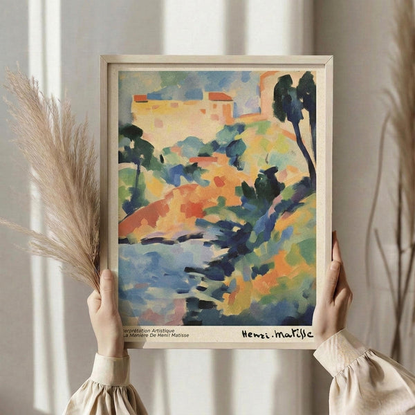 May include: Person in beige ruffled sleeves holding framed Henri Matisse landscape print featuring vibrant watercolor-style brushstrokes in blues, oranges, greens, and yellows depicting rolling hills, trees, and buildings. Dried wheat stalks visible on left side against soft neutral curtained background.