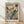 May include: Framed art print leaning against white wall on light wooden floor with natural sunlight casting shadows. The artwork shows a colorful Fauvism-style landscape painting with vibrant blues, oranges, greens, and yellows depicting rolling hills, trees, and buildings. Features loose, expressive brushwork in watercolor or oil paint style with French text at bottom and artist signature. Natural wood frame with white matting.