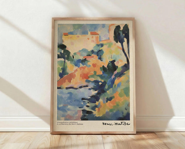 May include: Framed art print leaning against white wall on light wooden floor with natural sunlight casting shadows. The artwork shows a colorful Fauvism-style landscape painting with vibrant blues, oranges, greens, and yellows depicting rolling hills, trees, and buildings. Features loose, expressive brushwork in watercolor or oil paint style with French text at bottom and artist signature. Natural wood frame with white matting.