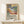 May include: Framed art print featuring a colorful tropical landscape with palm trees, foliage, and water in Matisse's distinctive Fauvist style using vibrant blues, greens, oranges, and pinks. Below the artwork is text reading "Matisse: A Master of Color and Form" with exhibition details. The print sits in a thin wooden frame on a light wood surface next to a small white gift box, displayed against a cream wall.